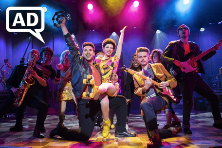 Beautiful_AudioDescribed_AD Production photograph from Beautiful - The Carole King Musical. Little Eva (Amena El-Kindy) smiles as she sits on the knee of a musician (Kevin Yates). The pair are smiling with an arm aloft, surrounded by other musicians and studio workers including Gerry Goffin (Tom Milner). Eva is wearing a 70s patterned dress and yellow roller skates, with cropped black hair, beneath candy-coloured lights in yellow, pink and blue. There is a white AD - Audio Description - logo top left.